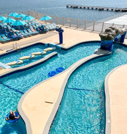 Aerial view of a pool area with blue water, lounge chairs, and a pier in the background on a sunny day.
