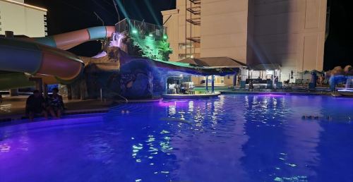 A brightly lit pool area at night, featuring a water slide and colorful lights reflecting on the water.