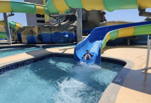 A child slides down a colorful water slide into a pool, creating splashes of water on a sunny day.