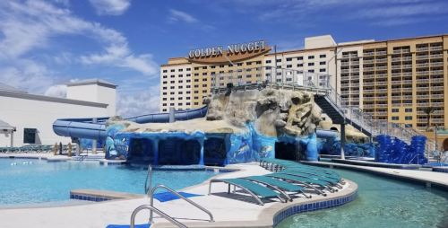 A resort pool area featuring a water slide, rock formations, and lounge chairs, with the Golden Nugget hotel in the background.