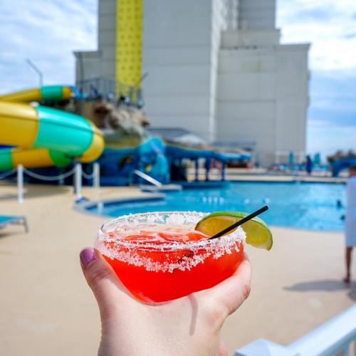 A hand holds a vibrant red cocktail with a lime, overlooking a pool and water slide on a sunny day.