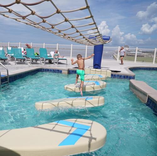 A child balances on floating platforms in a pool, with lounge chairs and people in the background under a blue sky.