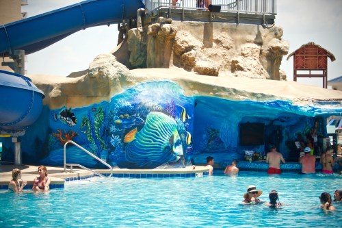 A vibrant pool scene featuring a water slide, colorful underwater mural, and people enjoying the water.