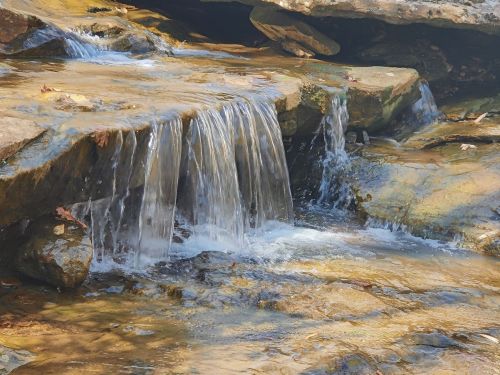 A small waterfall cascades over rocks into a clear stream, surrounded by natural scenery.
