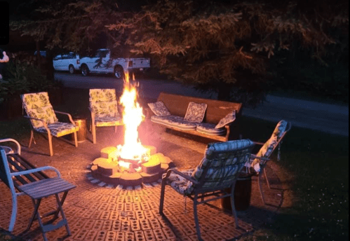 A cozy outdoor fire pit surrounded by chairs and a bench, with a bright fire and trees in the background at night.