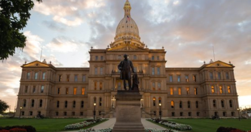 The Michigan State Capitol building at sunset, featuring a statue in the foreground and a dramatic sky.