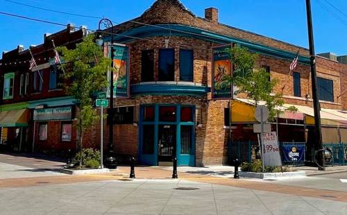A vibrant street corner featuring a brick building with colorful banners, trees, and nearby shops under a clear blue sky.