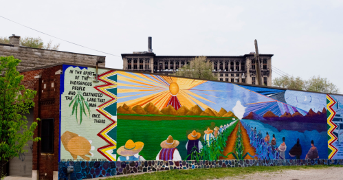 Colorful mural depicting indigenous people, sun, and landscapes, with a historic building in the background.