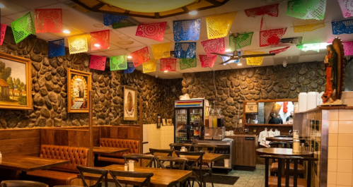 Cozy restaurant interior with stone walls, wooden tables, and colorful papel picado decorations hanging from the ceiling.