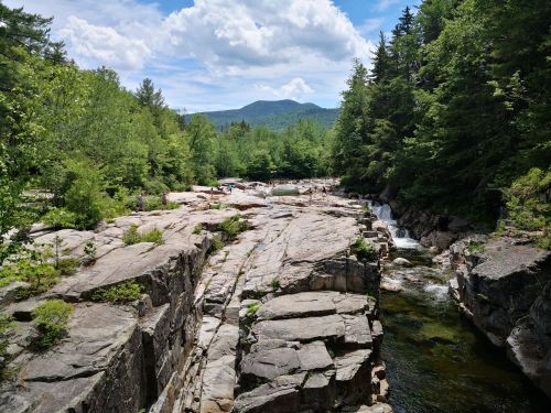 A scenic view of rocky terrain beside a river, surrounded by lush greenery and mountains under a partly cloudy sky.