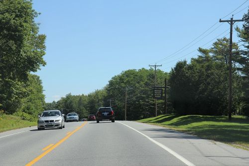 A two-lane road with cars driving, surrounded by green trees and clear blue sky. Power lines are visible along the roadside.