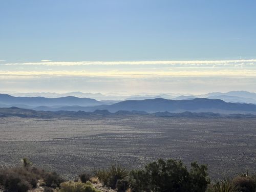 A vast desert landscape with layered mountains in the distance under a clear blue sky and wispy clouds.