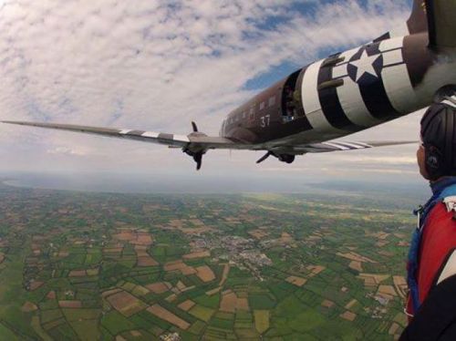A skydiver looks up at a vintage military plane flying over a patchwork of green fields and blue skies.