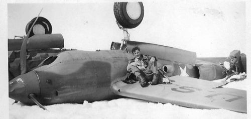 A pilot sits on the wing of a crashed aircraft in a snowy landscape, with another person working nearby.