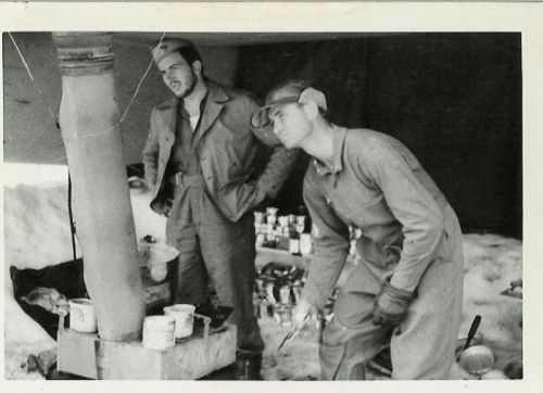 Two soldiers in military attire working near a stove in a snowy environment, surrounded by supplies and equipment.