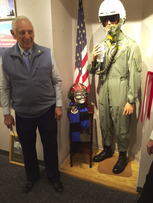 An elderly man stands next to a mannequin in a flight suit, with an American flag in the background.