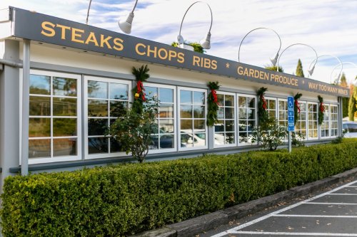 Exterior of a restaurant with a sign reading "Steaks, Chops, Ribs, Garden Produce, Way Too Many Wines," decorated for the holidays.