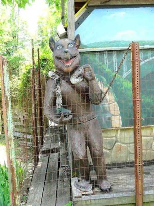 A whimsical, life-sized statue of a dog holding a snake, standing in a fenced area with greenery in the background.