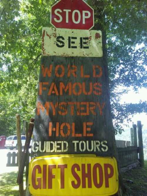 A weathered sign on a tree advertising a "World Famous Mystery Hole" with guided tours and a gift shop.