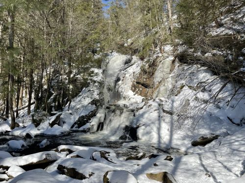 A snowy landscape featuring a waterfall cascading over rocks, surrounded by trees in a winter setting.