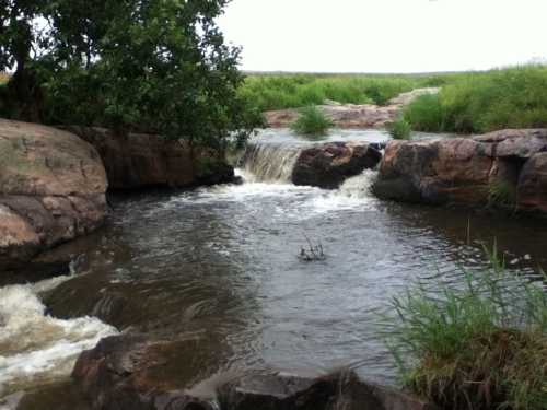 A serene river flows over rocks, creating a small waterfall, surrounded by lush greenery and a cloudy sky.