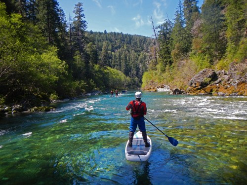 A person paddleboarding on a clear river surrounded by lush greenery and rocky banks under a blue sky.