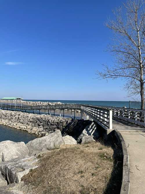 A serene waterfront scene with a wooden walkway, rocky shore, and clear blue sky.