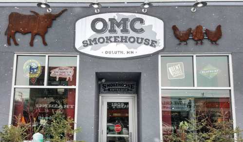 Exterior of OMC Smokehouse in Duluth, MN, featuring a sign and decorative metal animal silhouettes. Snow is falling.