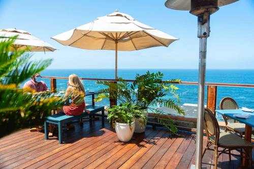 A couple sits at a table under an umbrella on a deck overlooking the ocean, surrounded by greenery.