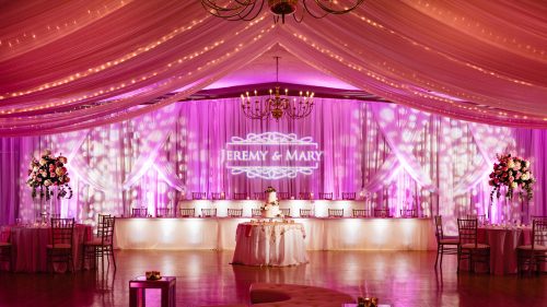 Elegant wedding reception with pink lighting, floral arrangements, and a backdrop featuring "Jeremy & Mary."