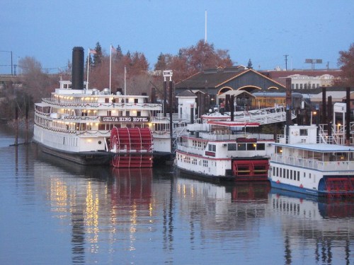 A riverboat hotel docked alongside smaller boats, reflecting in calm water with trees in the background.