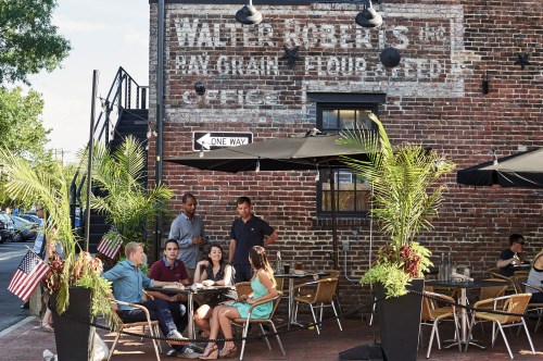A group of people dining outdoors at a café with a vintage brick wall and greenery, enjoying a sunny day.
