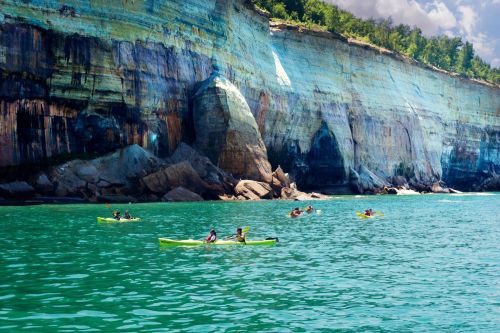 Kayakers paddle in turquoise waters near colorful cliffs under a bright sky.