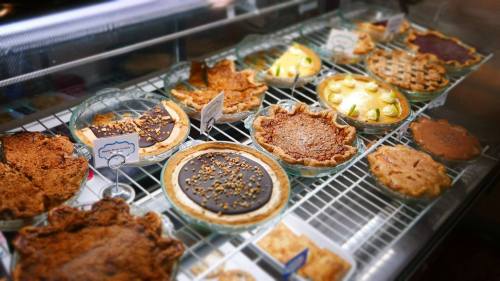 A display of various pies in glass dishes, including chocolate, fruit, and nut varieties, on a wire rack.