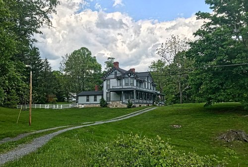 A large, historic house with a wraparound porch, surrounded by green grass and trees under a cloudy sky.