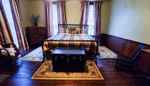 Cozy bedroom featuring a metal bed, patterned bedding, wooden furniture, and large windows with striped curtains.