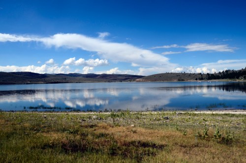 A serene lake reflecting clouds and mountains under a clear blue sky, surrounded by grassy shores.