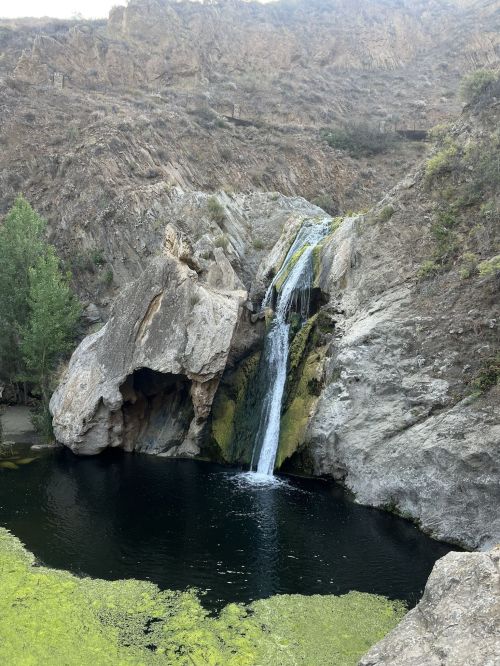A serene waterfall cascading into a dark pool, surrounded by rocky cliffs and green vegetation.