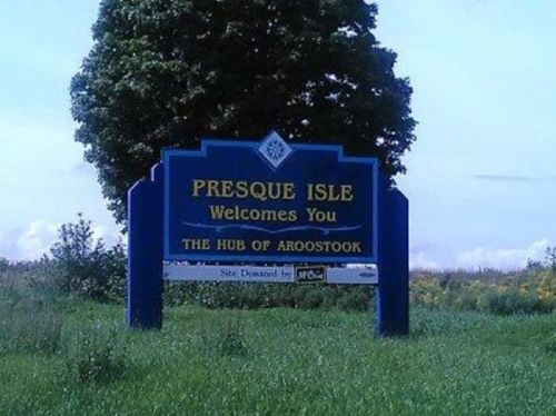 Sign welcoming visitors to Presque Isle, stating "The Hub of Aroostook," surrounded by green grass and trees.