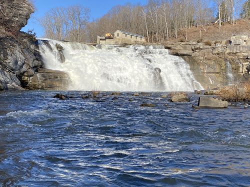 A waterfall cascades over rocky cliffs, with a building visible in the background and trees lining the shore.