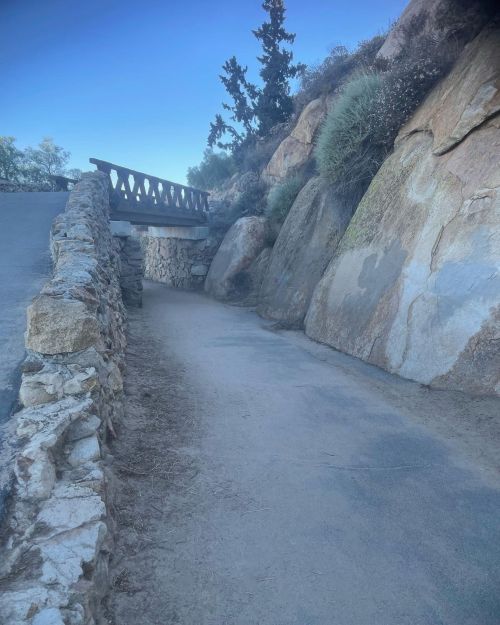 A winding path leads to a wooden bridge, flanked by rocky terrain and greenery under a clear blue sky.