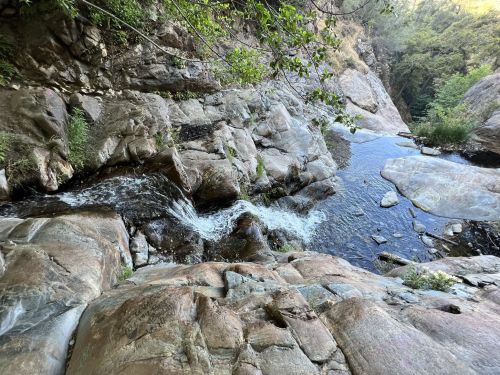 A serene river flows over smooth rocks, surrounded by lush greenery and steep canyon walls.