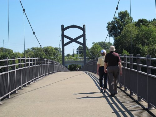 A couple walks hand in hand on a suspension bridge surrounded by greenery under a clear blue sky.