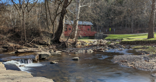 A serene stream flows past a red covered bridge, surrounded by bare trees and a grassy area in a peaceful landscape.