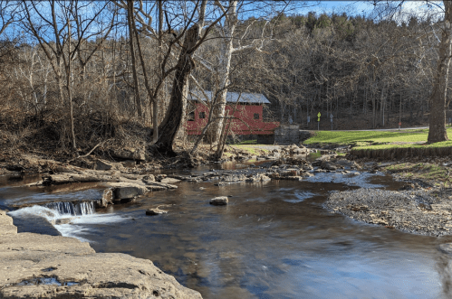 A serene stream flows past a red barn, surrounded by bare trees and a green hillside in early spring.