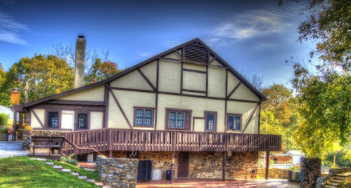 A charming two-story house with a wooden deck, stone foundation, and a sloped roof, surrounded by trees and blue sky.