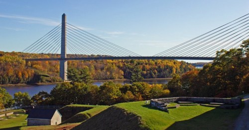 A cable-stayed bridge spans a river, surrounded by autumn foliage and green hills under a clear blue sky.
