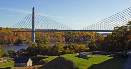 A cable-stayed bridge spans a river, surrounded by autumn foliage and green hills under a clear blue sky.