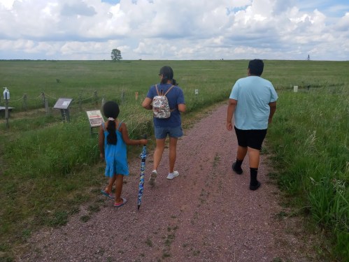 Three children walking on a gravel path through a grassy field, with clouds in the sky and signs along the trail.