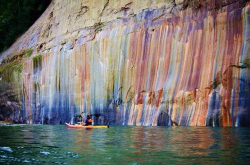 A kayaker paddles near a colorful, striped rock formation along the water's edge.